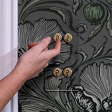 Hand operating a clear antique brass 4G vertical two-way toggle switch on a floral-patterned wall.
