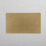 Antique Brass 2G Blank Plate for Sockets & Modules with rounded corners on a white textured wall.