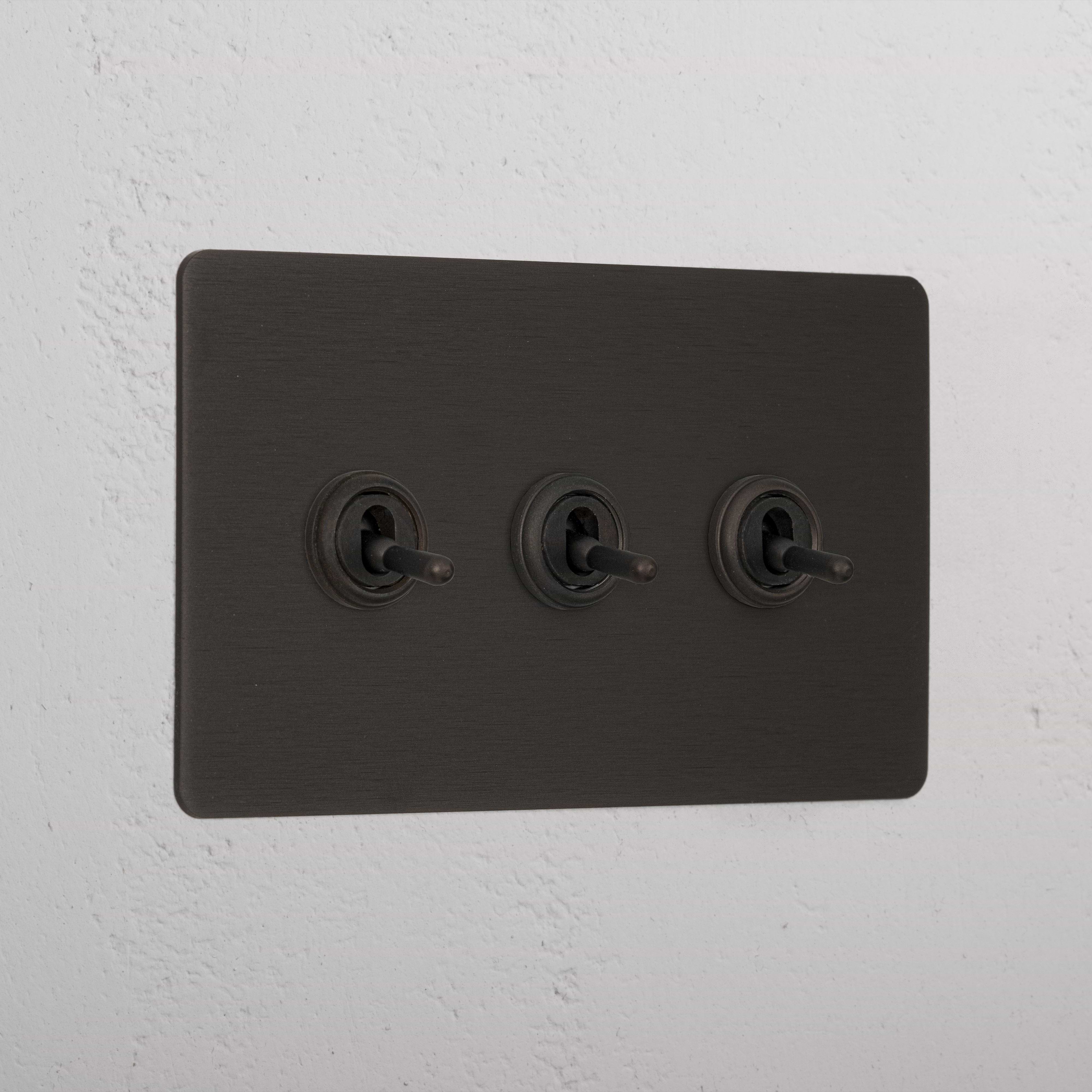 Bronze panel with three retractive toggle switches (3G) mounted on a white wall.