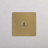 Antique brass 1G centre retractive toggle switch on a white textured wall.