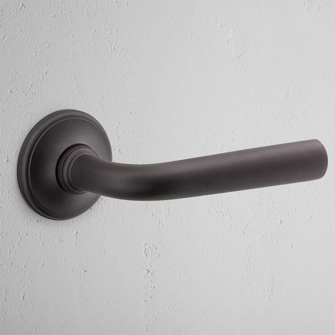 Apsley Unsprung Door Handle in bronze with a sleek black finish against a textured white wall.