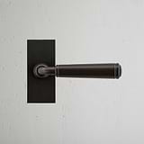 Digby Short Plate Fixed Door Handle in bronze on a white textured wall.