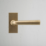 Apsley Short Plate Sprung Door Handle in antique brass on a textured white wall, offering an elegant look.