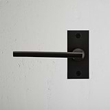 Clayton Short Plate Sprung Door Handle in bronze, mounted on a textured white wall for an elegant finish.