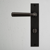 Digby Long Plate Sprung Door Handle & Thumbturn in bronze, displayed on a white textured wall.