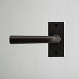 Apsley Short Plate Sprung Door Handle in bronze on a textured white wall.