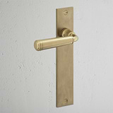 Digby Long Plate Sprung Door Handle in Antique Brass on a textured white wall, perfect for classic decor.