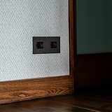 Bronze black switched fused spur (FCU) isolator switch on a patterned wall with wooden trim and flooring.