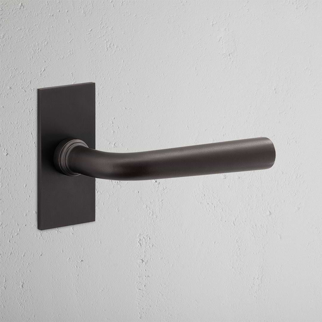 Apsley Short Plate Fixed Door Handle - Bronze, elegantly displayed on a textured white wall.