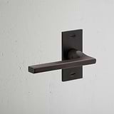 Clayton Short Plate Sprung Door Handle in bronze finish mounted on a white textured wall.