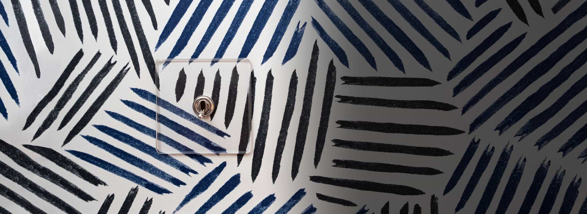 Durchsichtiger Rahmen mit vernickeltem Kipphebelschalter auf weißer Tapete mit schwarzen und blauen Strichen