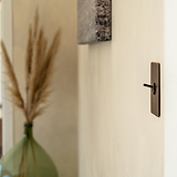 Kipphebelschalter – Bronze – an eine cremefarbene Wand montiert, grüne Glasvase im Hintergrund. 