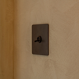 Kipphebelschalter 1x in Einfachrahmen – Bronze – an eine cremefarbene Wand montiert, aus der rechten Perspektive fotografiert. 