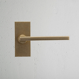 Manija de puerta de latón antiguo Clayton con placa corta y mecanismo de resorte sobre pared blanca texturizada.