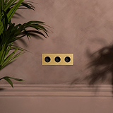 Enchufe de pared triple de latón antiguo con insertos negros, Módulo redondo Schuko x3, sobre pared marrón.