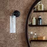 Aplique de pared Claremont pequeño en bronce con vidrio acanalado, uno a cada lado de un espejo circular en la pared de un cuarto de baño. 