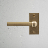 Manilla de puerta Harper con placa corta en latón antiguo sobre pared de textura ligera, diseño elegante y atemporal.