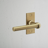 Manilla para puertas Harper con placa corta en latón antiguo sobre pared texturizada blanca.