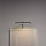 Una moderna lámpara de pared negra de la colección Harrow de 220 mm ilumina un marco vacío en una pared gris.