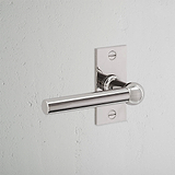 Manija de puerta Harper con placa corta y resorte en níquel pulido sobre pared texturizada blanca.