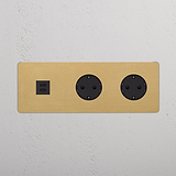 Marco triple con módulo USB redondo de 30W y dos bases Schuko, acabado latón antiguo con inserto negro.