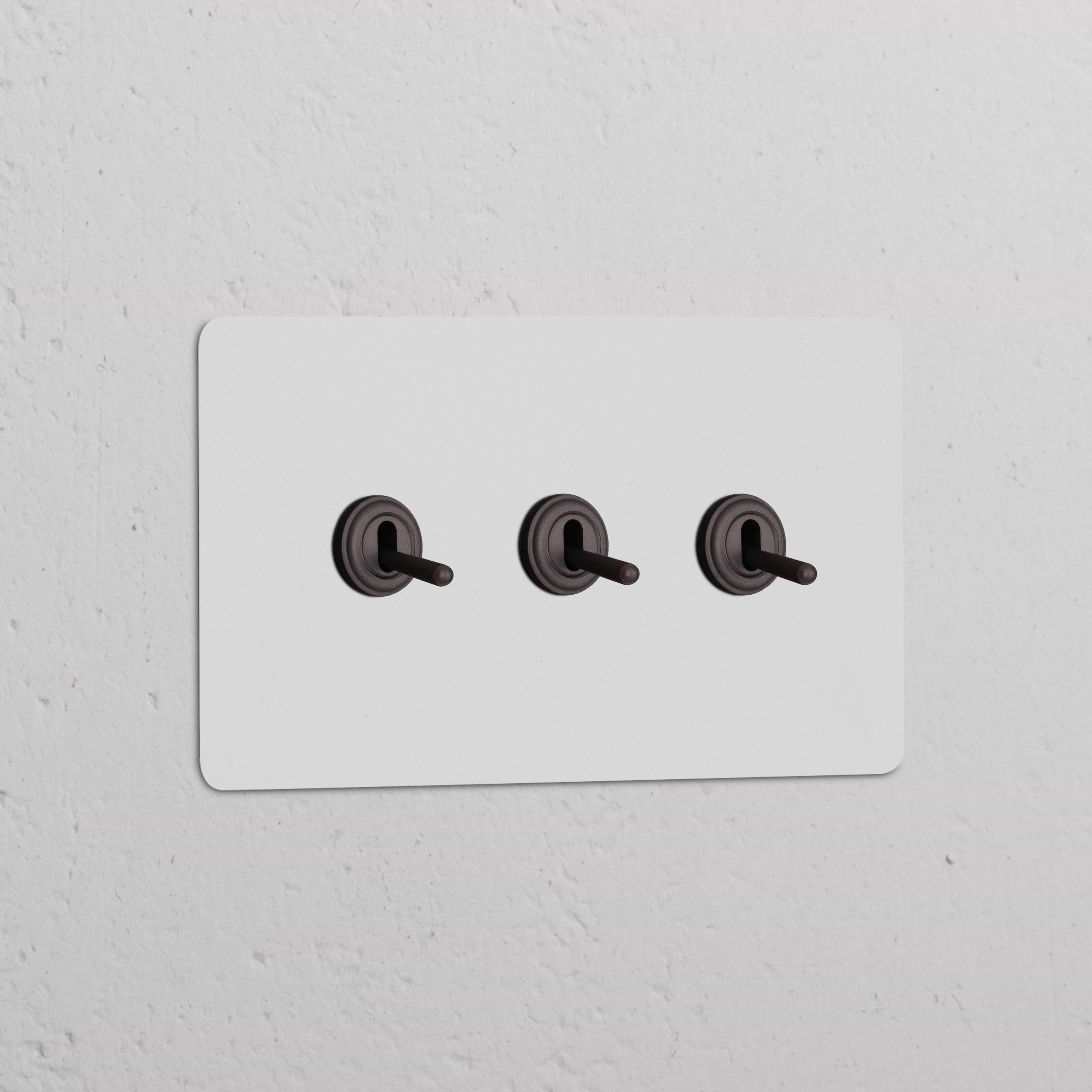 Interruptor conmutador de palanca x3 en marco doble pintable y bronce sobre pared blanca, elegante y funcional.