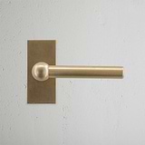Manilla para puertas Harper con placa corta de latón antiguo sobre pared gris texturizada.