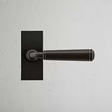 Poignée de Porte à Ressort à Plaque Courte Digby en bronze sur plaque rectangulaire, montée sur un mur blanc.