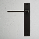 Clayton Poignée de Porte à Ressort à Plaque Longue - Bronze sur un mur texturé blanc, offrant une touche élégante.