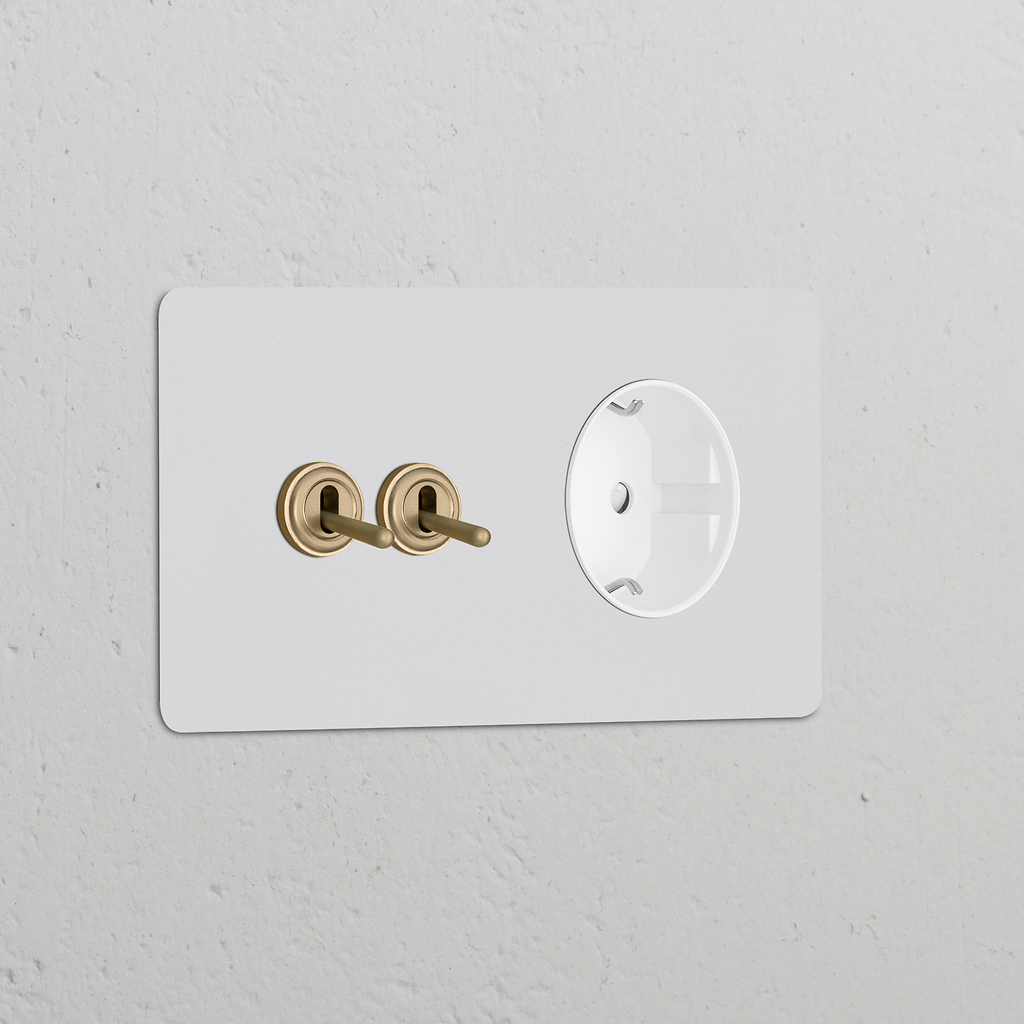 Doppio interruttore a levetta 2x con modulo Schuko Tipo F tondo, ottone antico bianco su parete bianca.