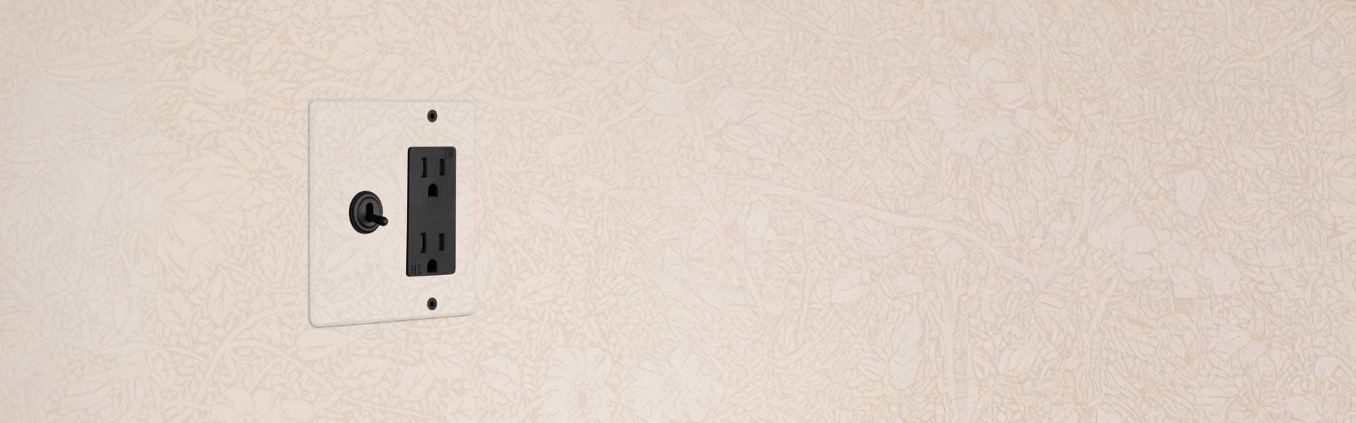 A black electrical outlet with a toggle switch installed on a beige wall with a subtle floral pattern.