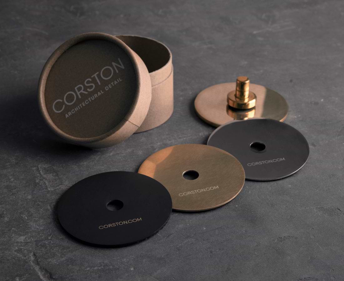 A round box labeled "CORSTON Architectural Detail" contains three decorative discs in black, bronze, and gold finishes, with a central attachment piece.
