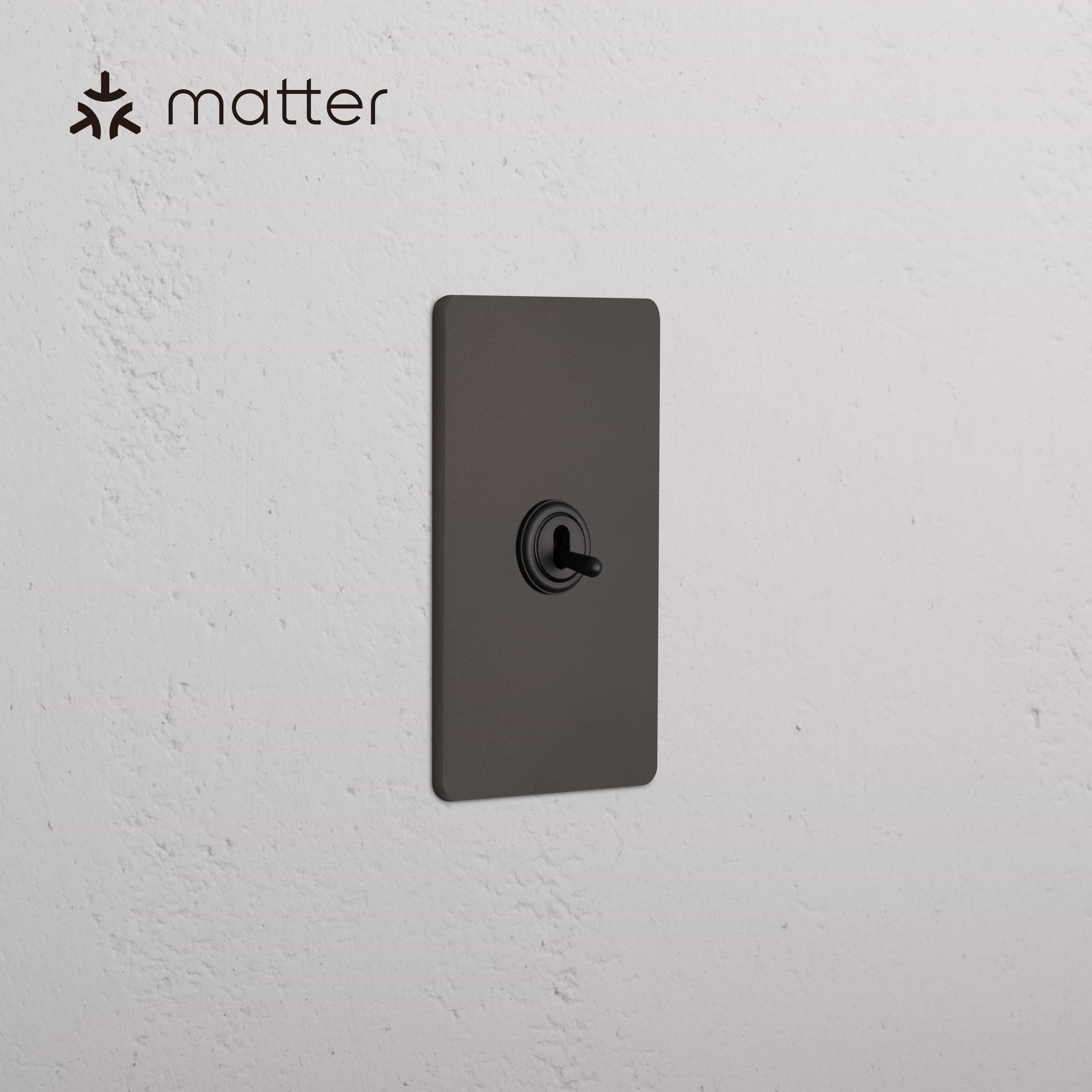 Grove 1G Smart Retractive Toggle Switch in bronze finish, shown mounted on a white textured wall.