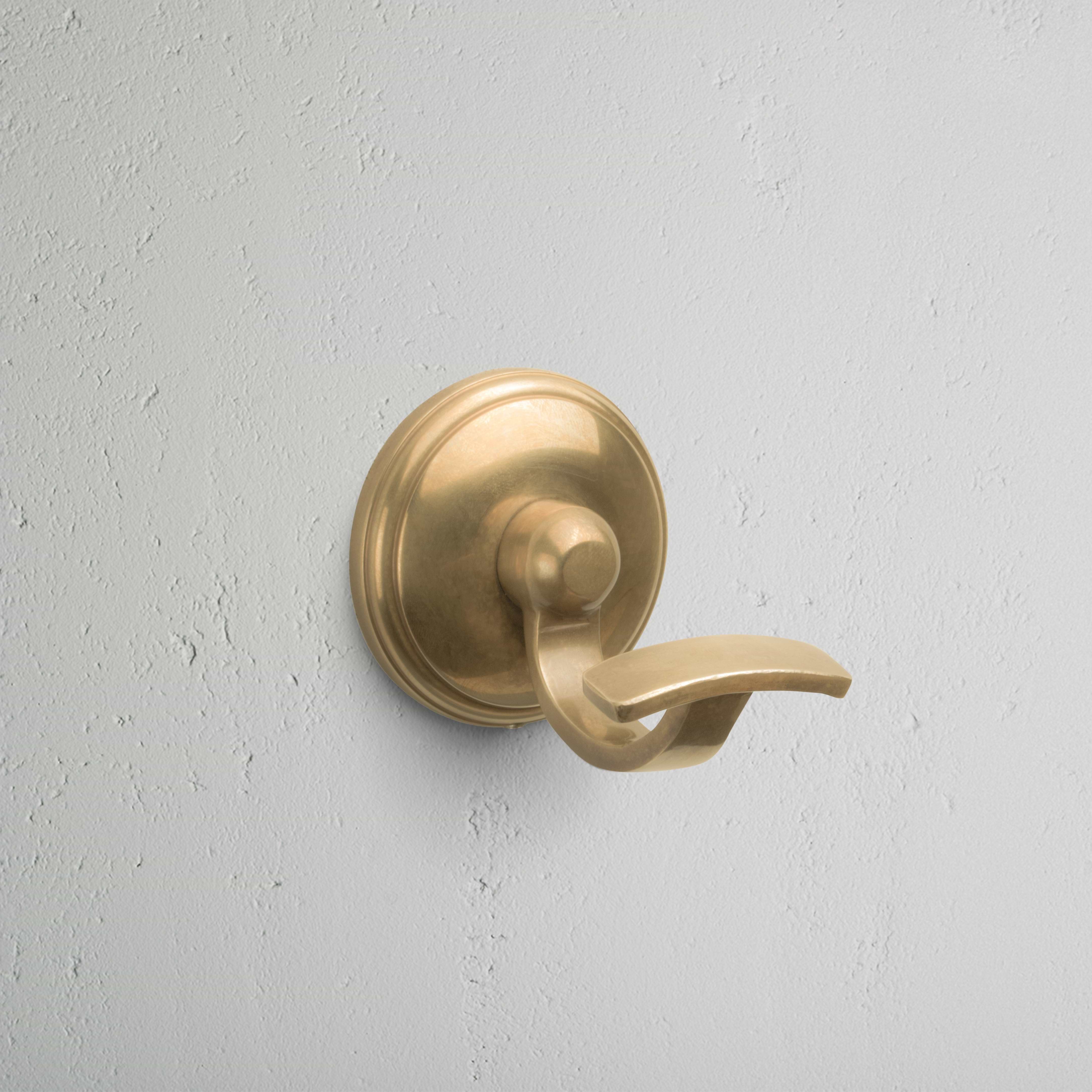 Antique brass Portland Hook with round base, mounted on a light gray textured wall.