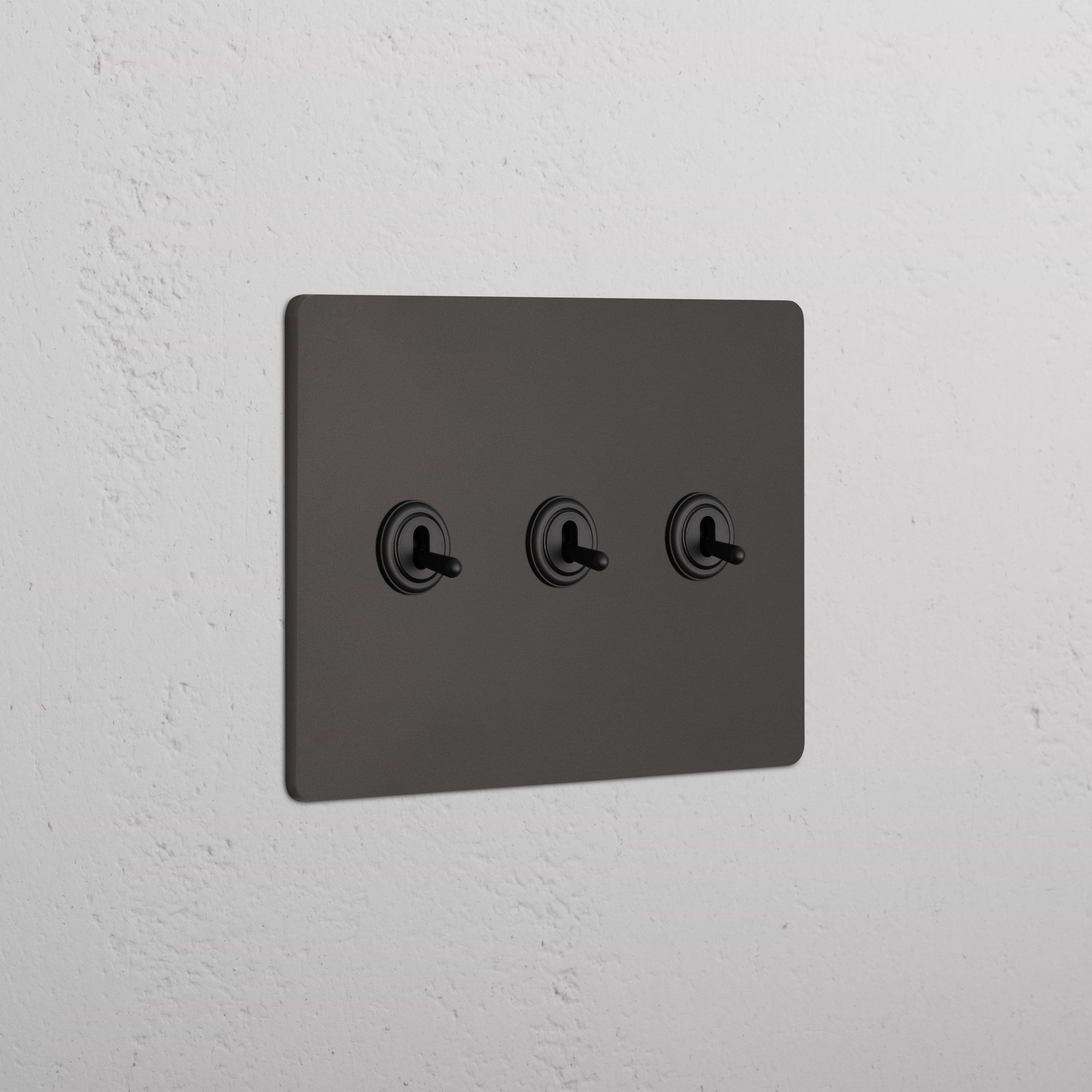 Grove 3G Toggle Switch in bronze with three toggle switches, installed on a white textured wall.