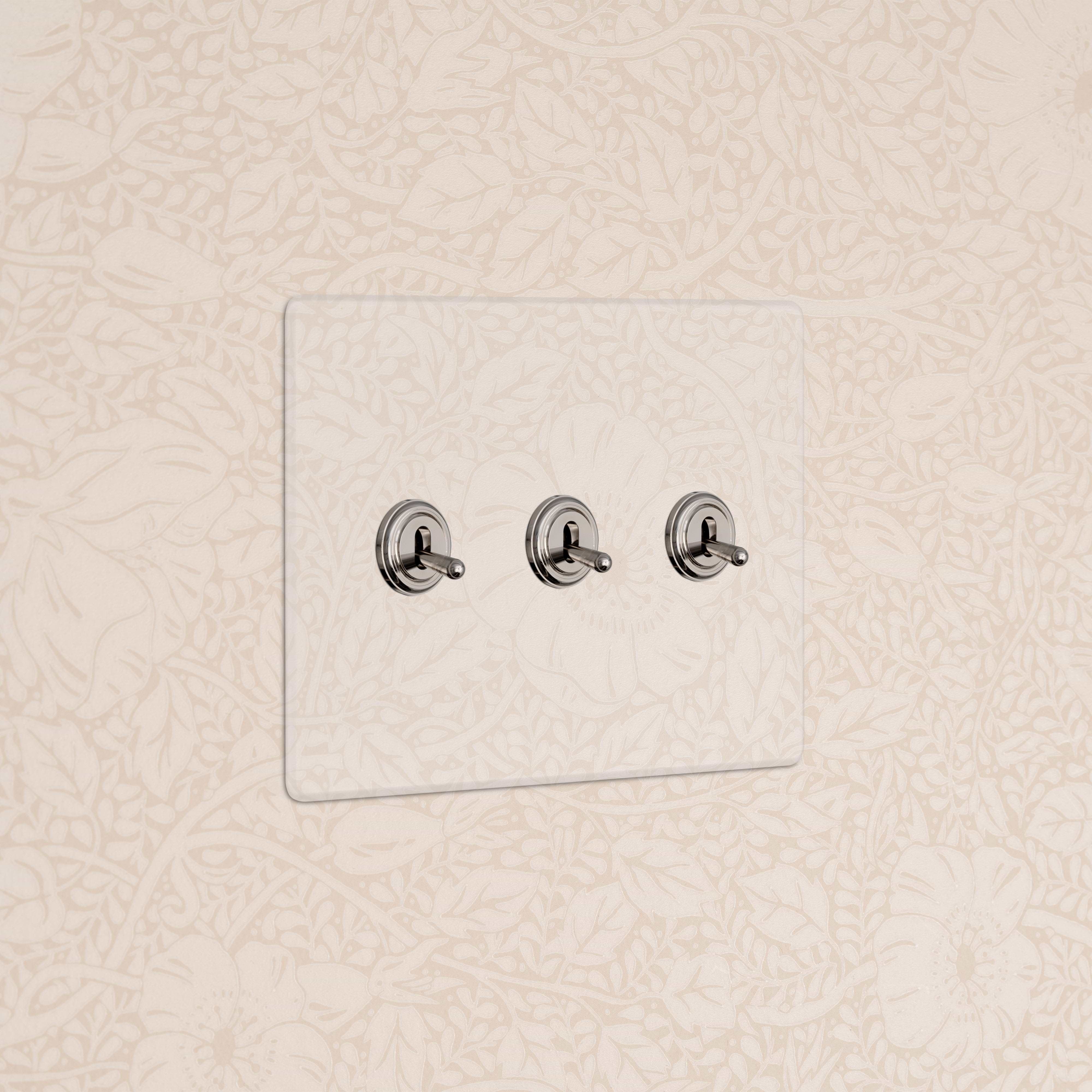 Kew 3G Toggle Switch in Clear Polished Nickel with three toggles, shown on a beige floral-patterned wall.