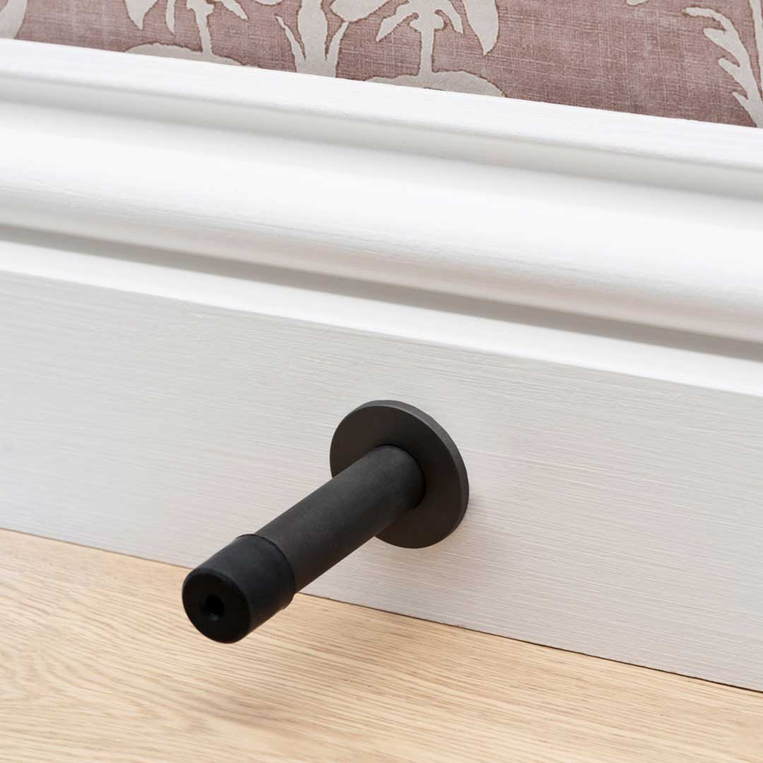 Montpelier Door Stop in bronze finish mounted on white baseboard above light wood floor, with patterned wallpaper.
