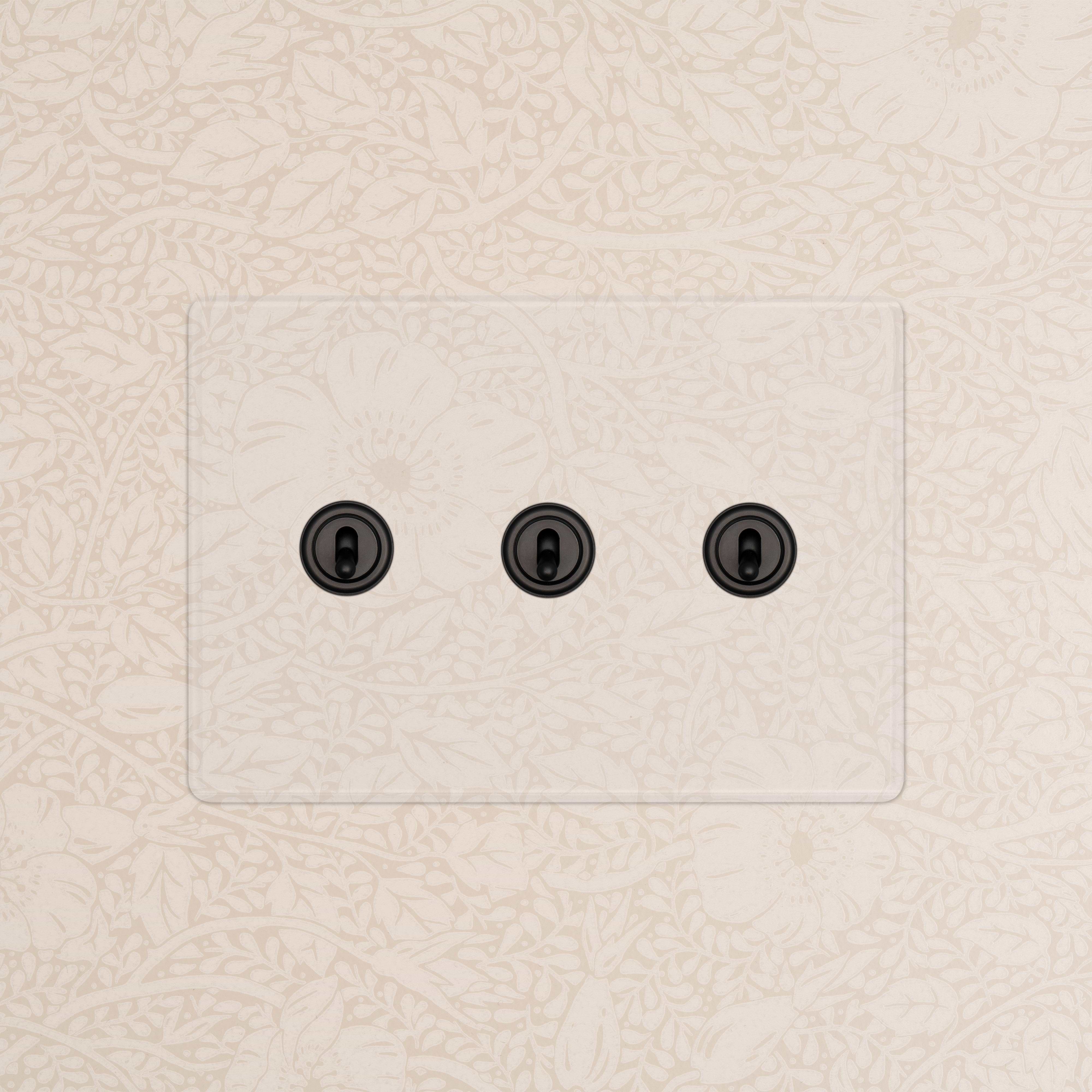 Kew 3G Toggle Switch in Clear Bronze finish with black toggles mounted on beige floral-patterned wallpaper.