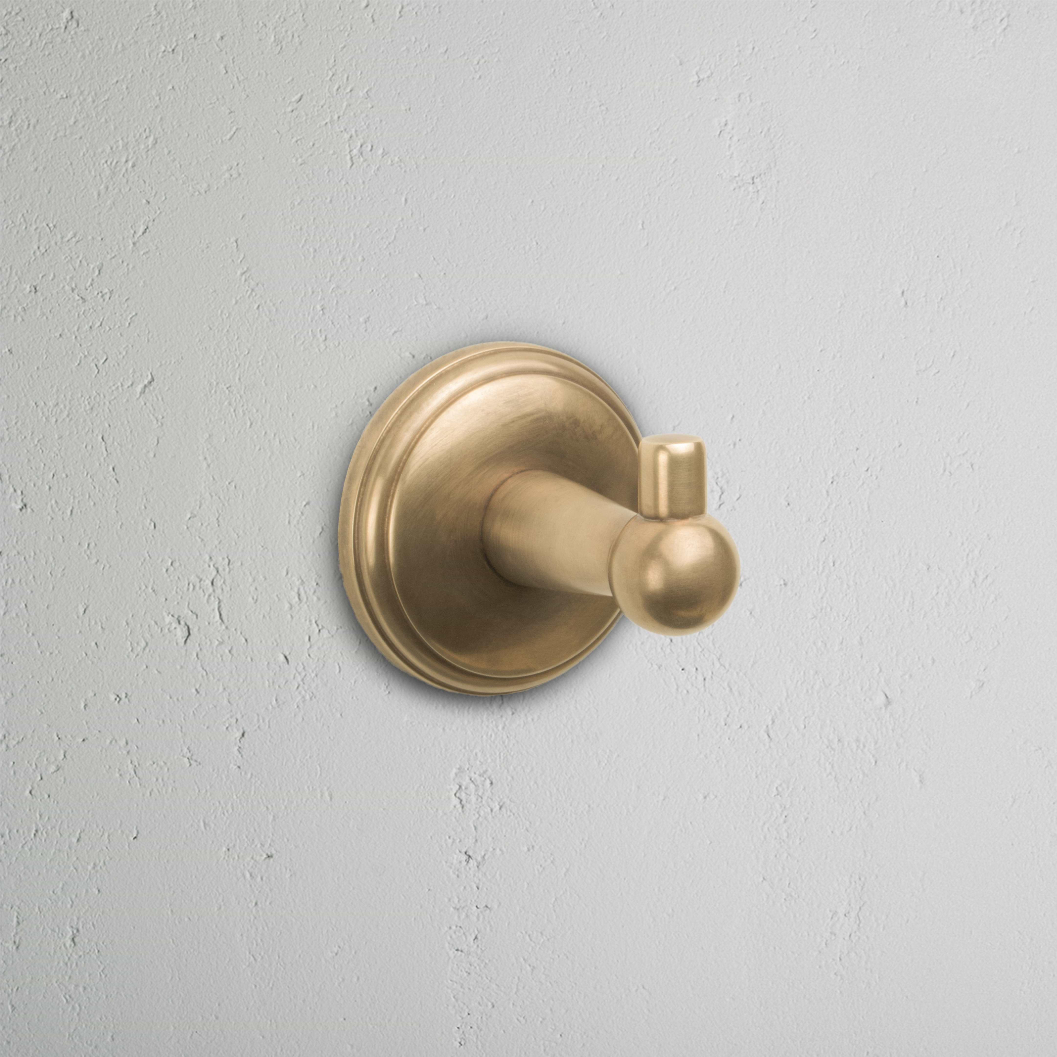 Hyde Hook in antique brass finish, with a round base, mounted on a light gray textured wall.