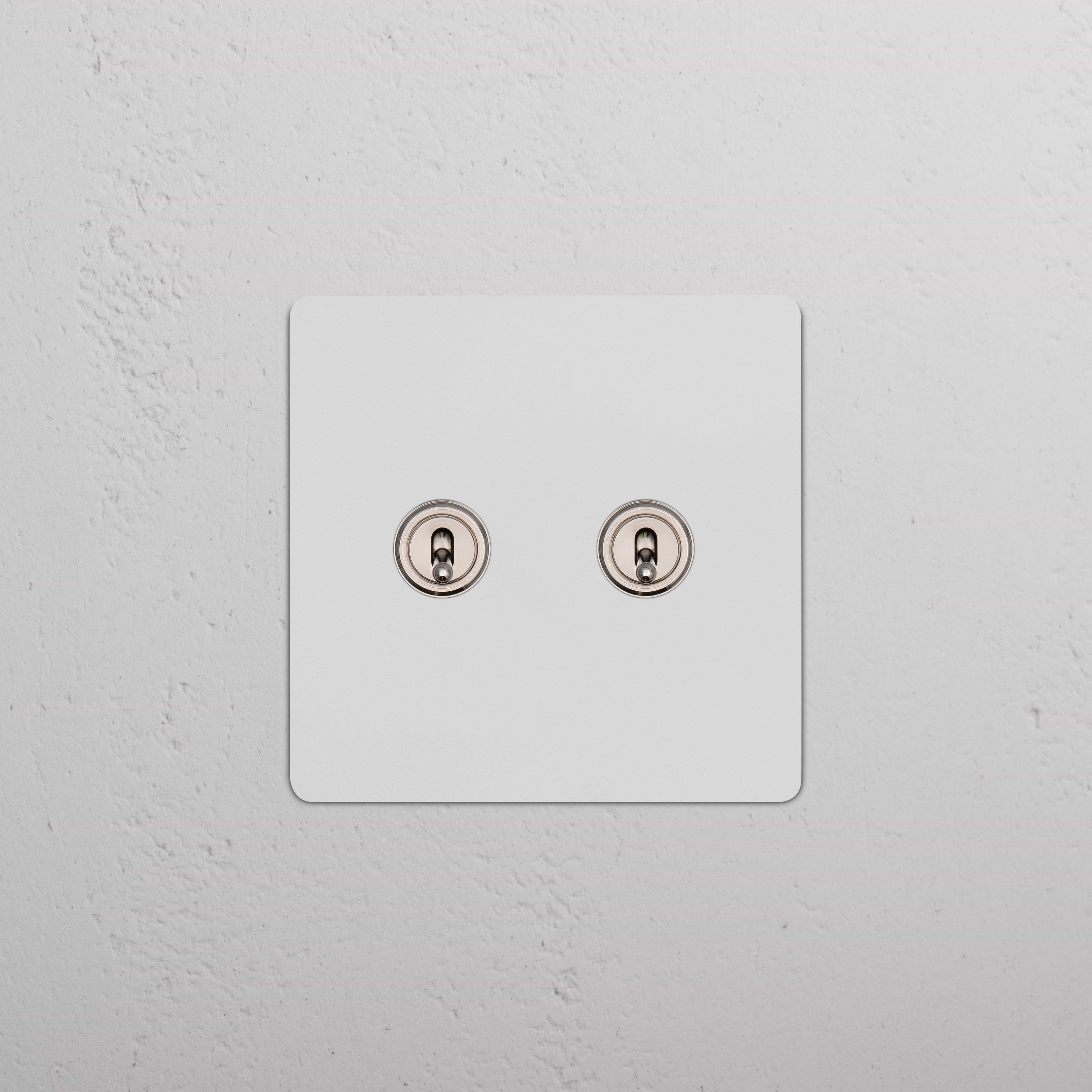 Kew 2G Toggle Switch in paintable polished nickel with two round toggles on a white textured wall.