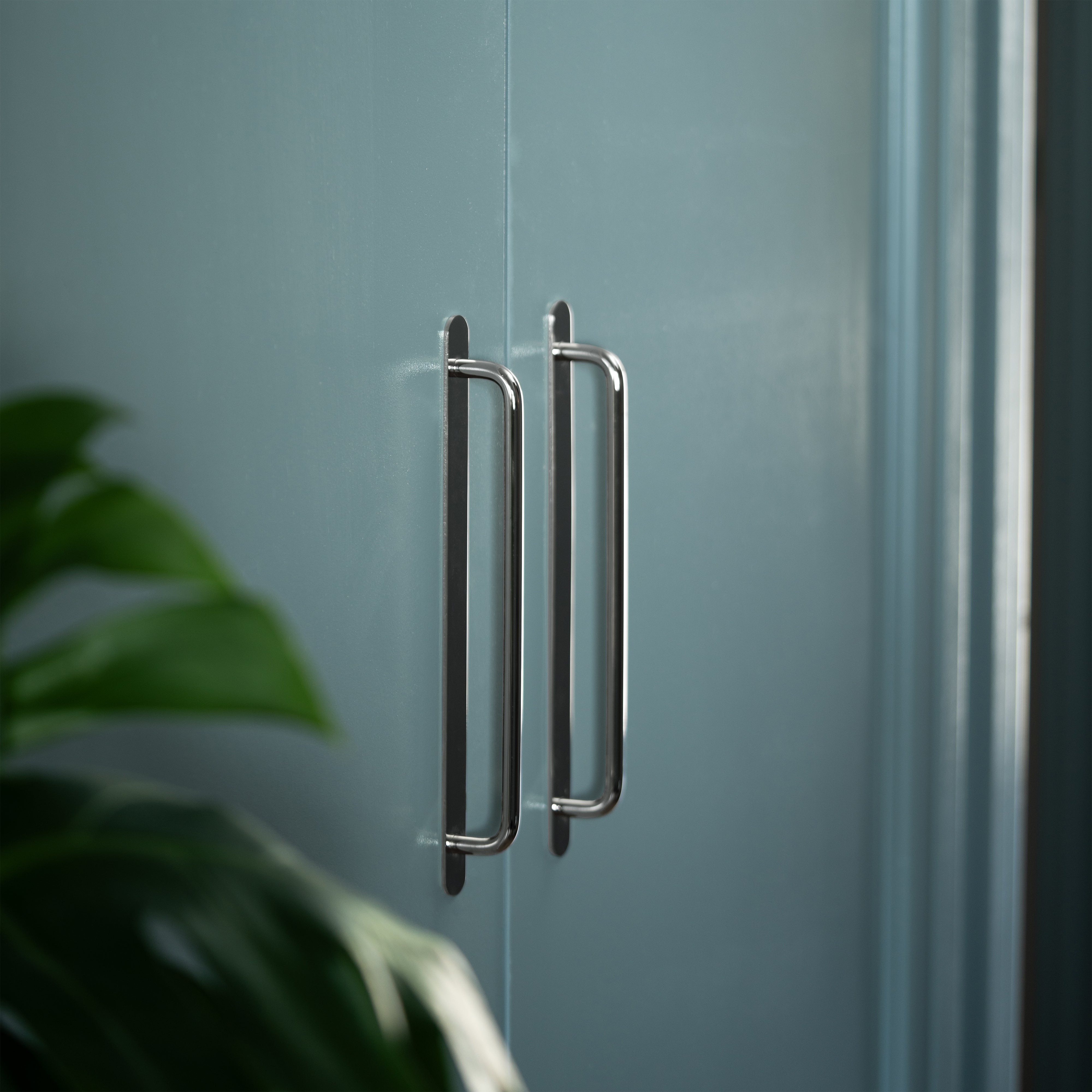 Kilburn Furniture Handle Medium in polished nickel, mounted on a blue cabinet door, with a green plant nearby.