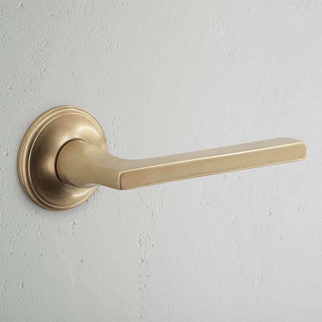 Clayton Passage Door Handle Set in antique brass with round base and straight lever on a light gray wall.