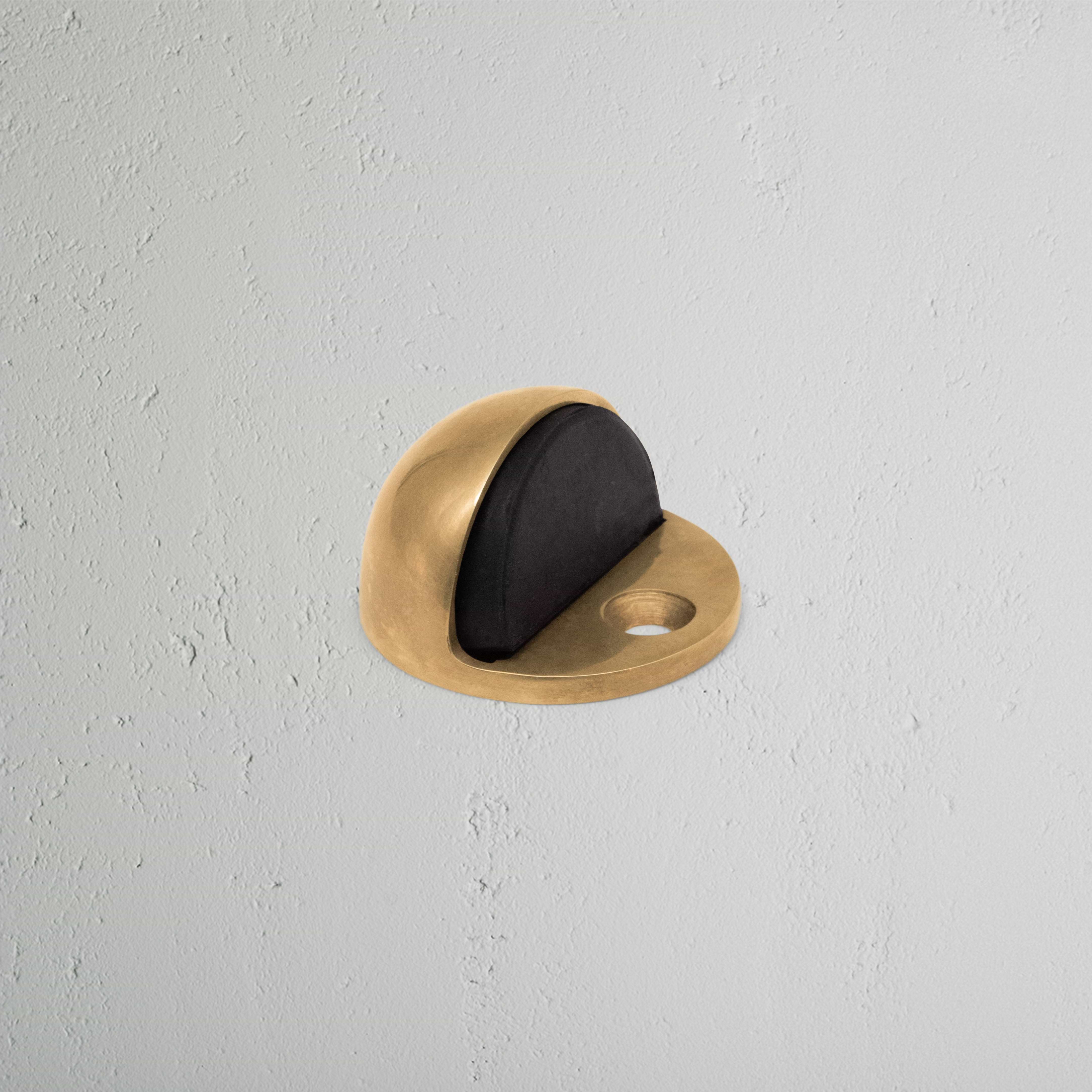 Clifton Half Moon Door Stop in antique brass with black rubber insert on a light gray textured wall.