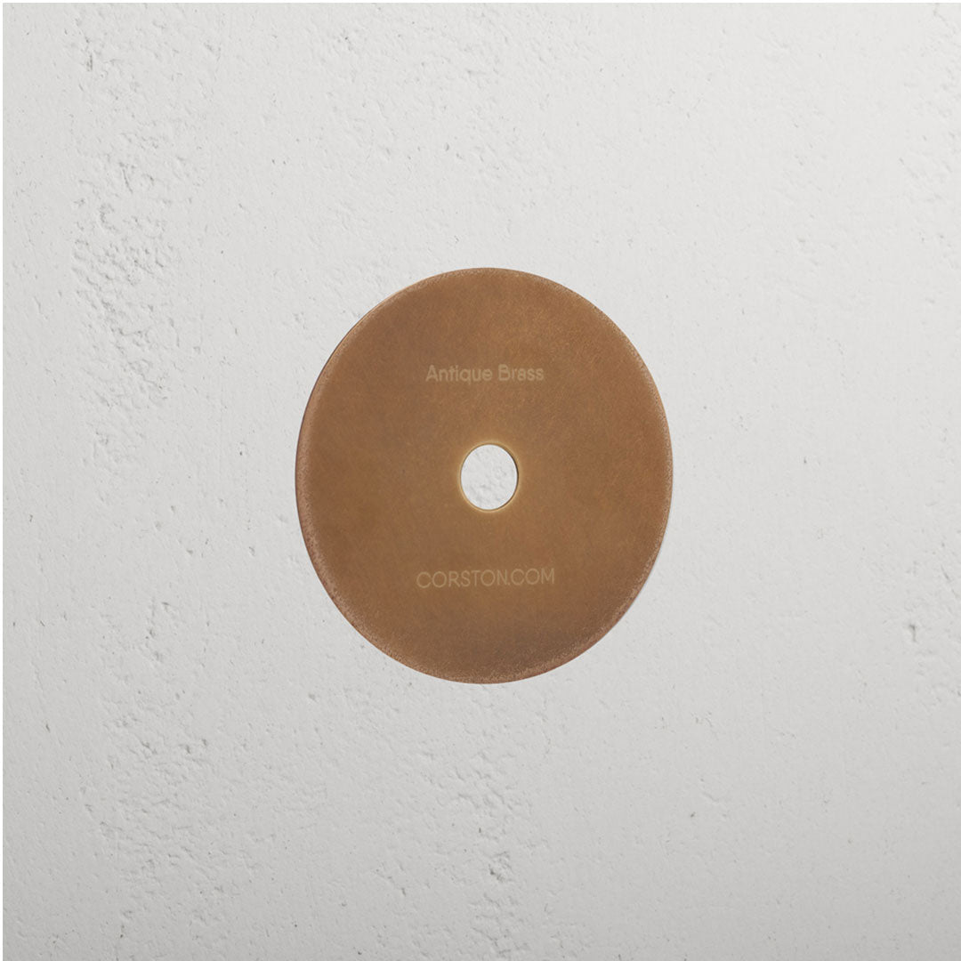 Circular Sample Finish in Antique Brass with central hole, mounted on white textured wall, labeled 'CORSTON.COM'.