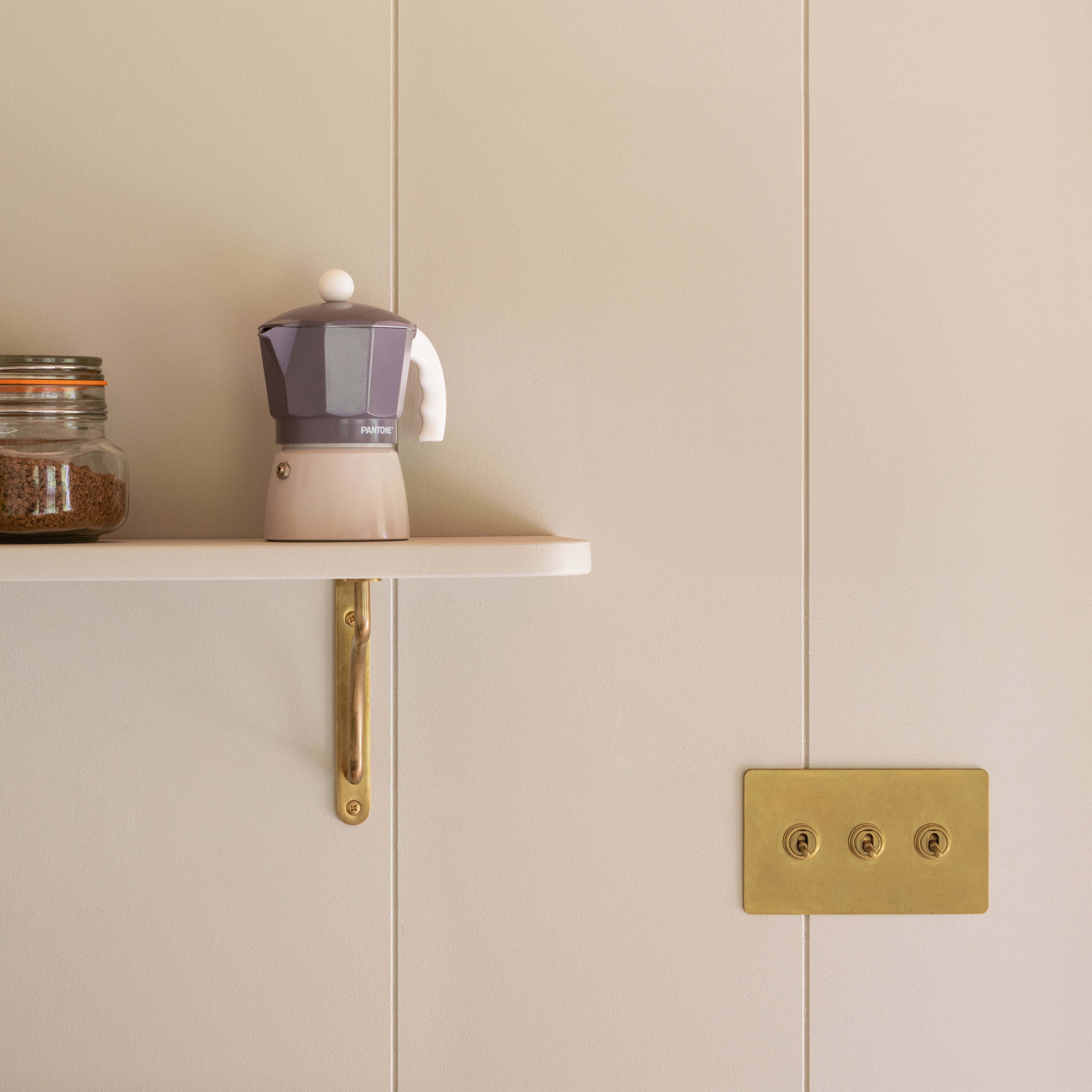 Small antique brass Kilburn shelf bracket supporting a white shelf against a beige wall.