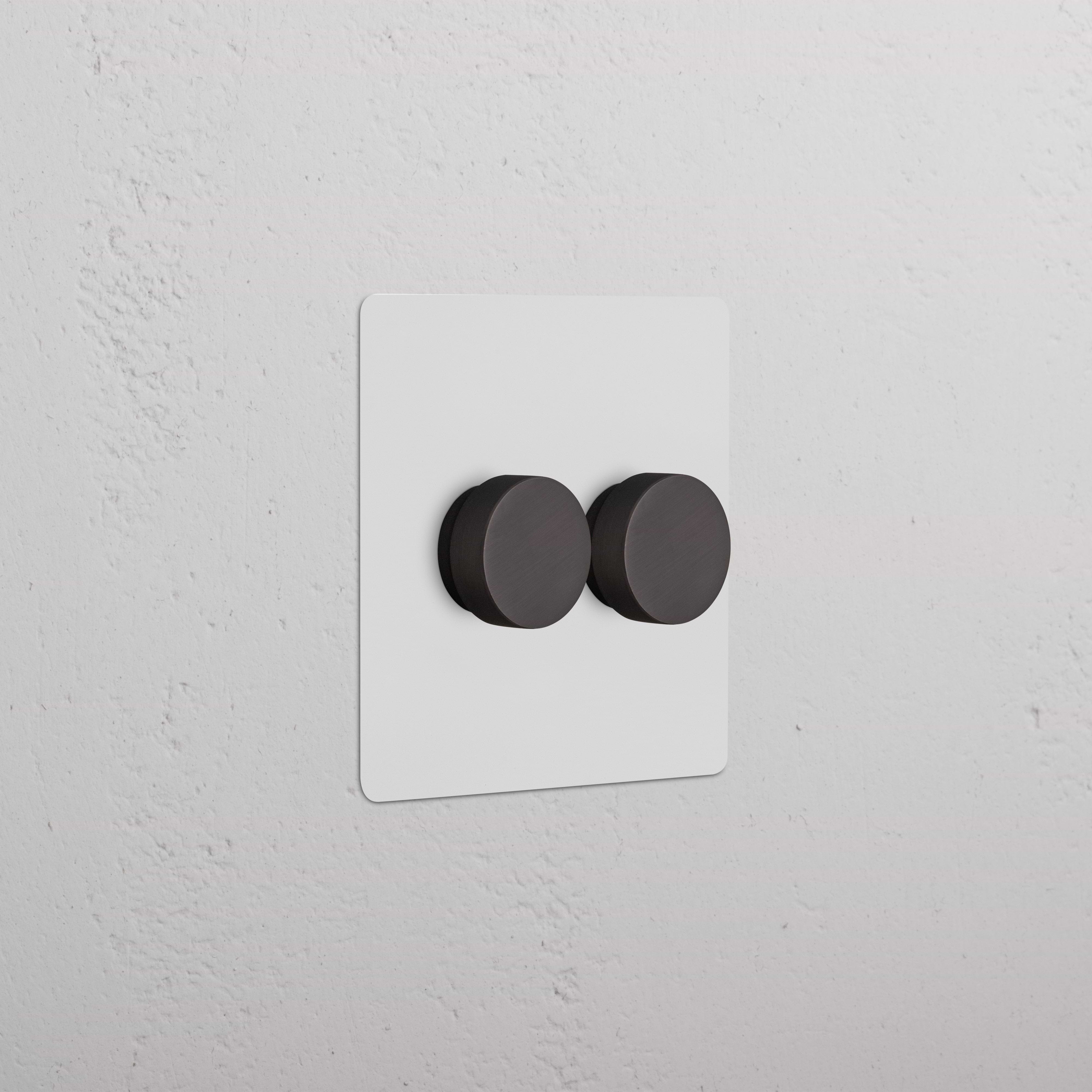 Kew 2G Dimmer Switch in paintable bronze with two round black knobs, shown on a wall.