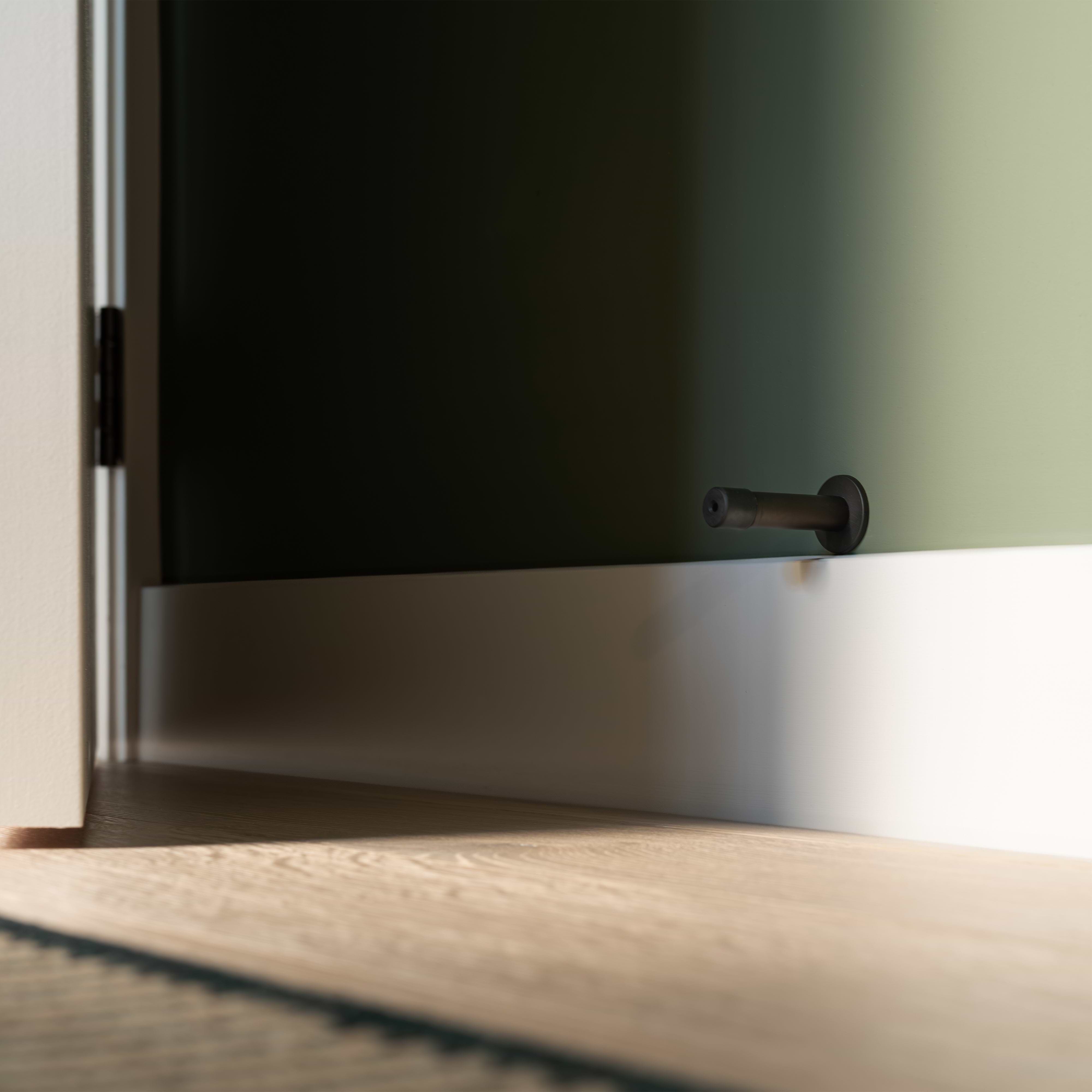 Bronze Montpelier Door Stop with a rubber tip mounted on a white baseboard in a sunlit room.