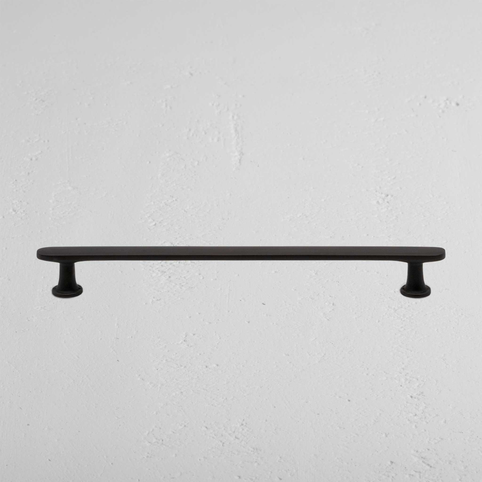 Milliner Furniture Handle Medium in bronze with rounded ends, mounted on a white textured wall.