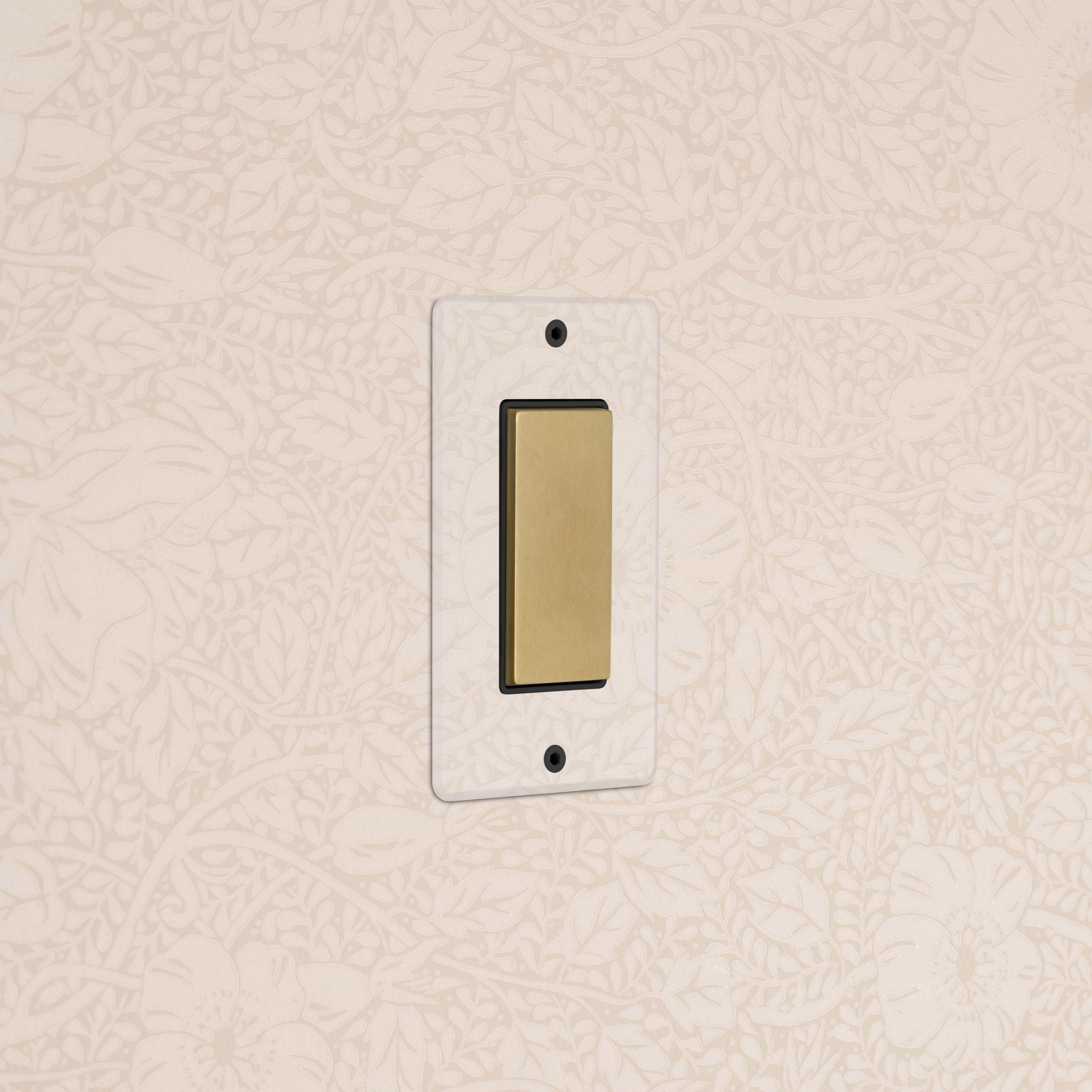 Kew 1G Center Retractive Rocker Switch in clear antique brass black on a wall with floral patterned wallpaper.