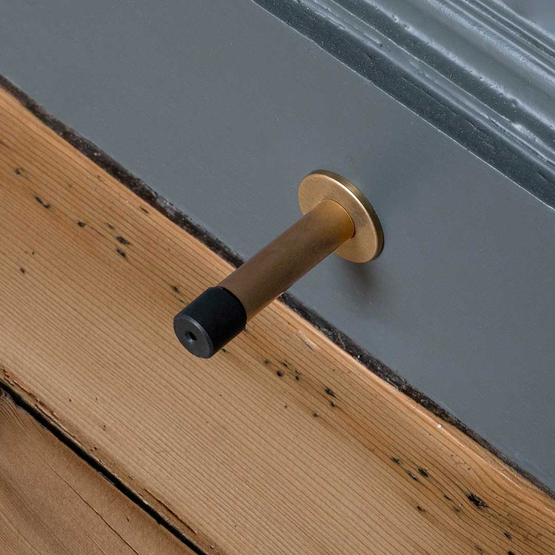 Montpelier Door Stop in Antique Brass with black rubber tip, mounted on a baseboard above a wooden floor.